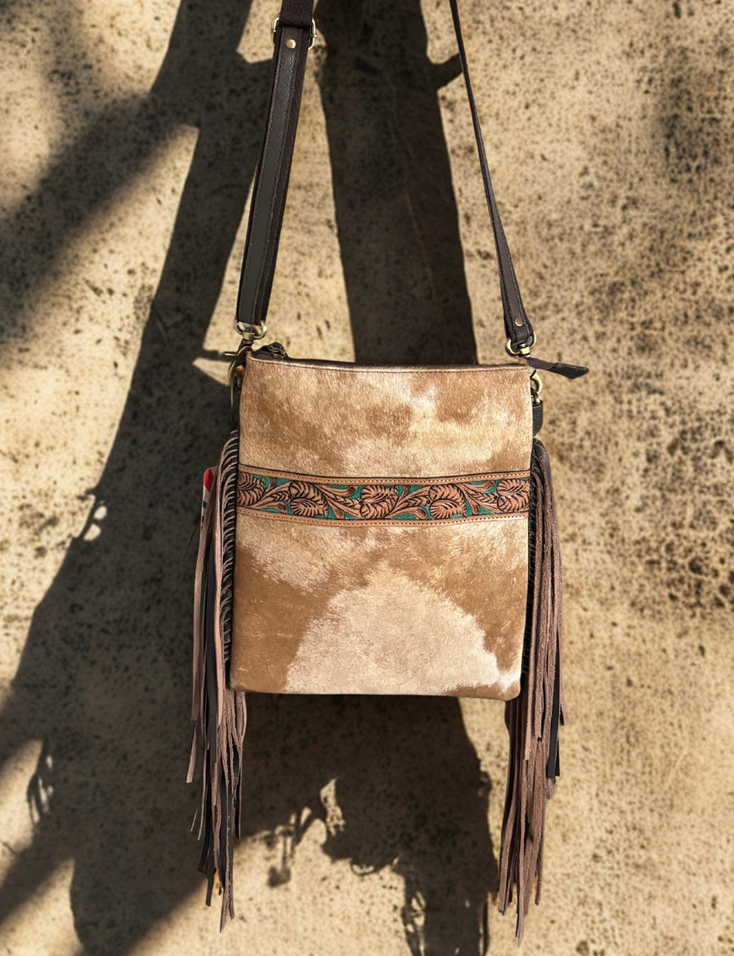 Beige leather crossbody bag with fringe and decorative strap on a textured surface